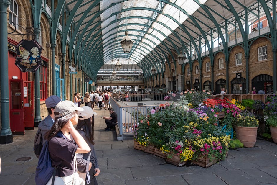 A large, arched glass and metal roof structure with a grid pattern, supported by teal-colored steel beams, extends over a covered walkway. Along the sides, hanging lantern-style lights provide illumin
